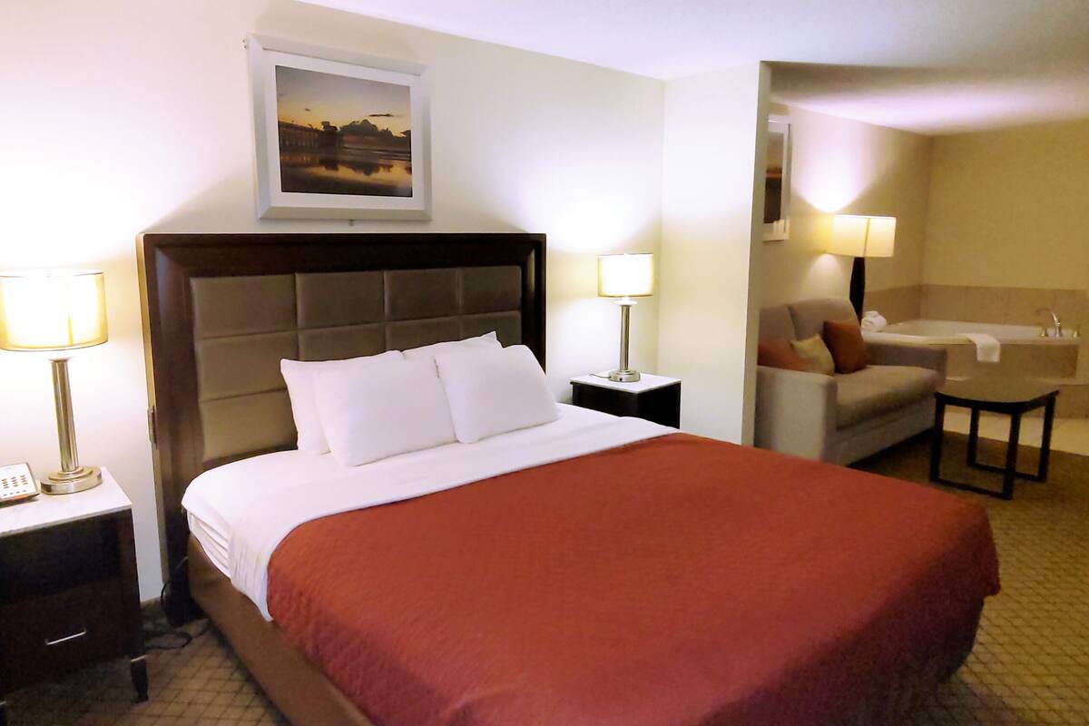 A hotel room with a large bed, two bedside tables with lamps, a sofa, a small table, and a bathroom visible through an open door.