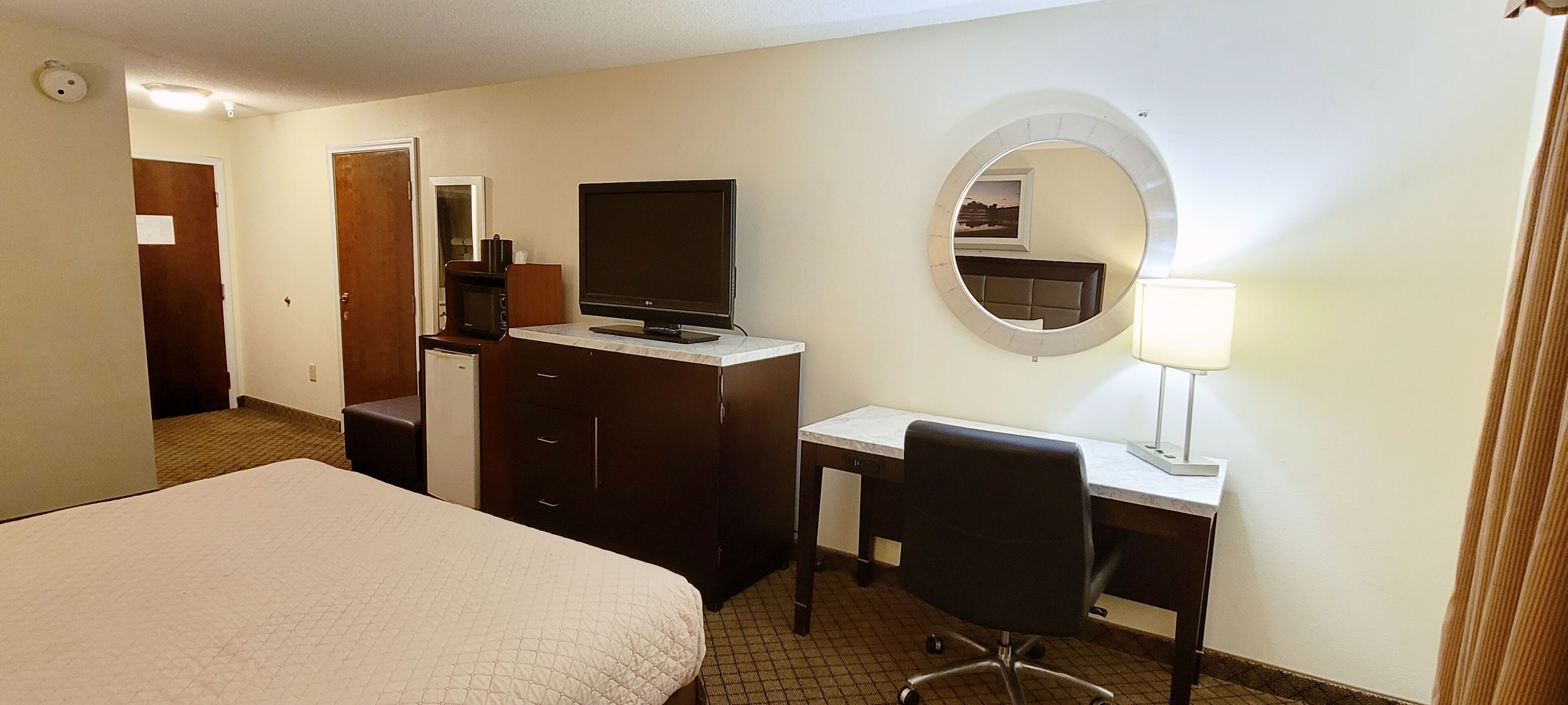 A hotel room with a bed, a desk with a chair, a television, a microwave, a dresser, and a mirror.