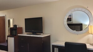 A hotel room with a flat-screen TV on a marble counter, a round mirror on the wall, a desk with a chair, and a small kitchenette with a microwave. - Home Inn & Suites (Montgomery)