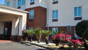A multi-story hotel with a beige facade, a sign reading "Home Innsuites," and a brick section on the lower level. There are palm trees and flowering bushes in the front yard. - Home Inn & Suites (Montgomery)