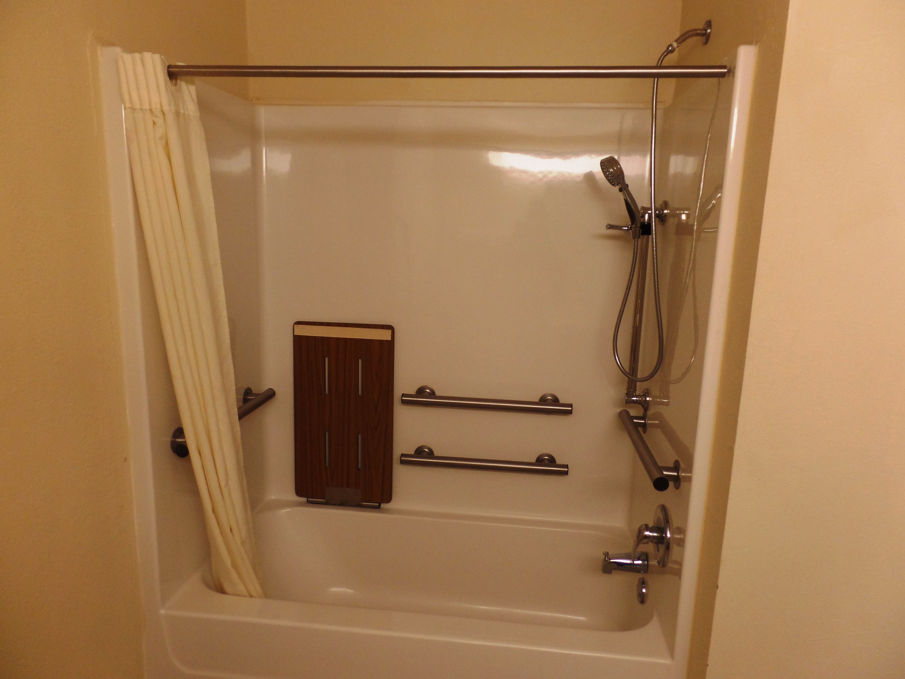 Une baignoire standard équipée d’un pommeau de douche, de barres d’appui et d’un panneau en bois.