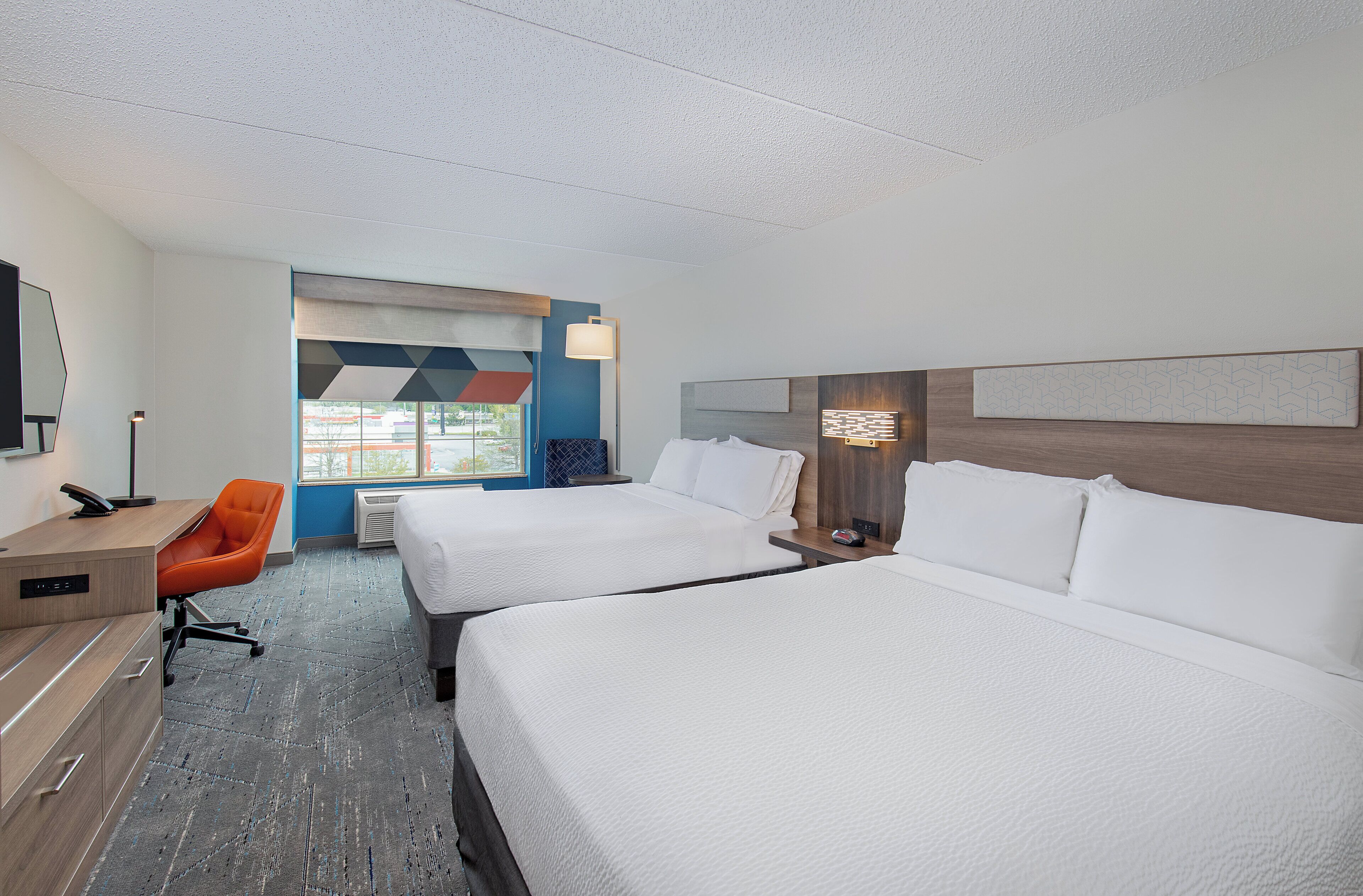 Una habitación de hotel con dos camas, un escritorio, una silla naranja y un ventanal grande.