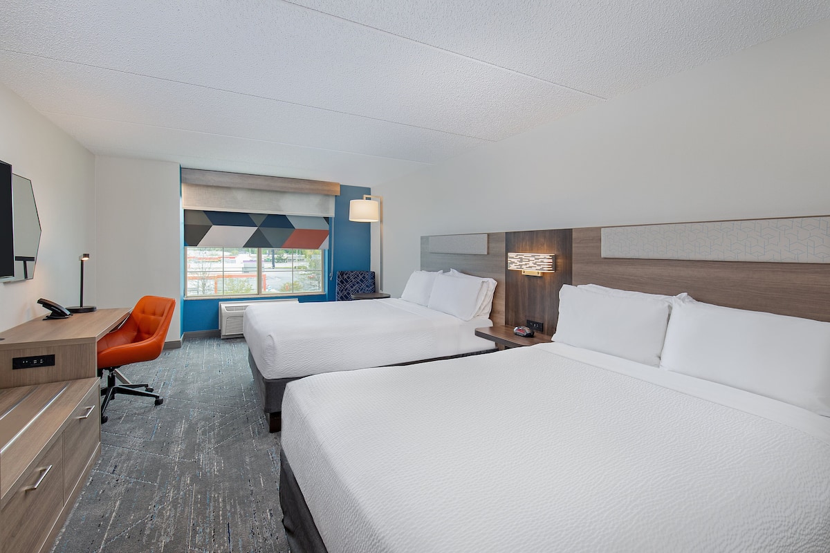 Ein Hotelzimmer mit zwei Betten, einem Schreibtisch, einem orangefarbenen Sessel und einem großen Fenster.