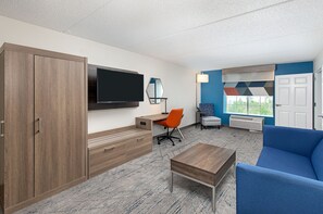 Een moderne hotelkamer met een houten garderobe, een flatscreen televisie, een bureau met een oranje bureaustoel, een blauwe bank en een groot raam met uitzicht op groen.