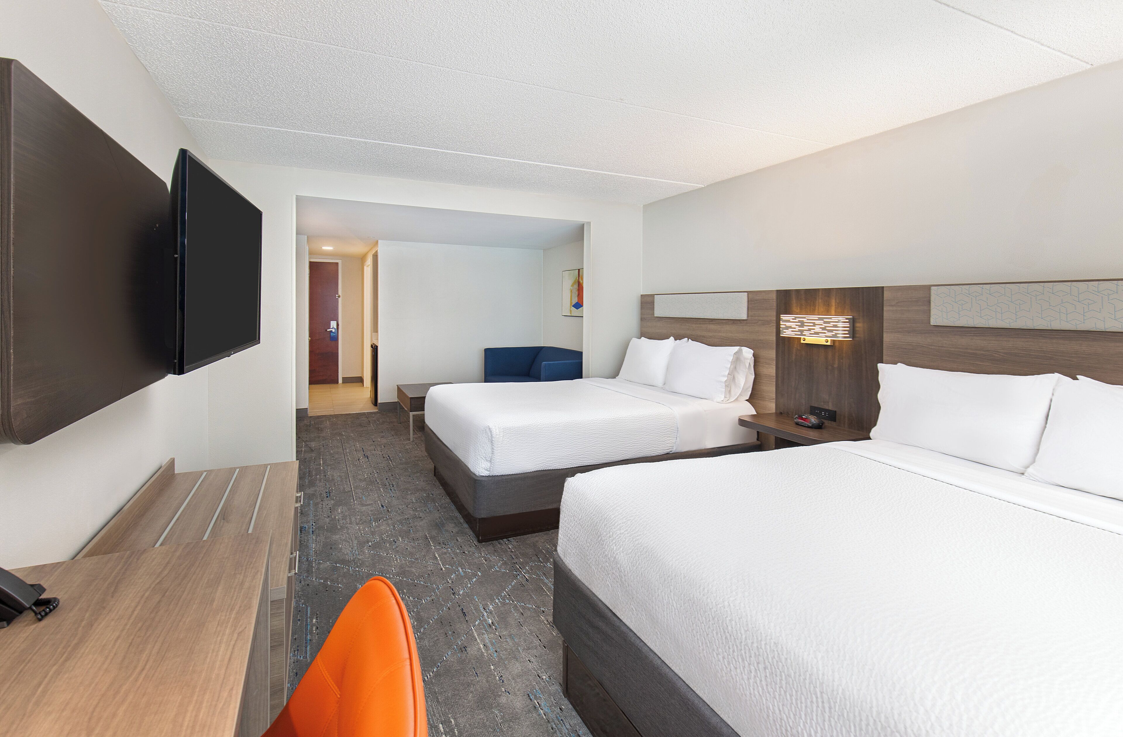 A hotel room with two beds, a desk, a chair, a flat-screen TV, and a door leading to a hallway.