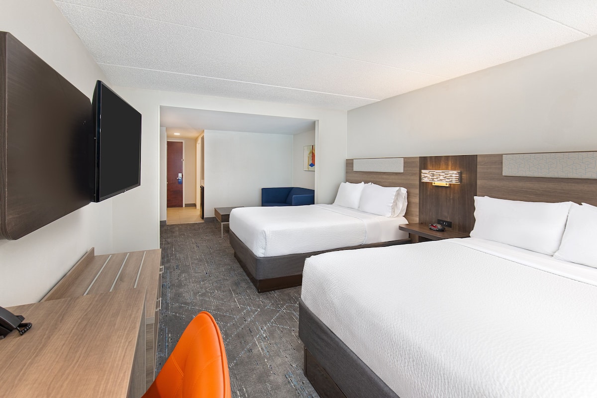 Ein Hotelzimmer mit zwei Betten, einem Schreibtisch, einem Stuhl, einem an der Wand montierten Fernseher und einer Tür zu einem weiteren Zimmer.