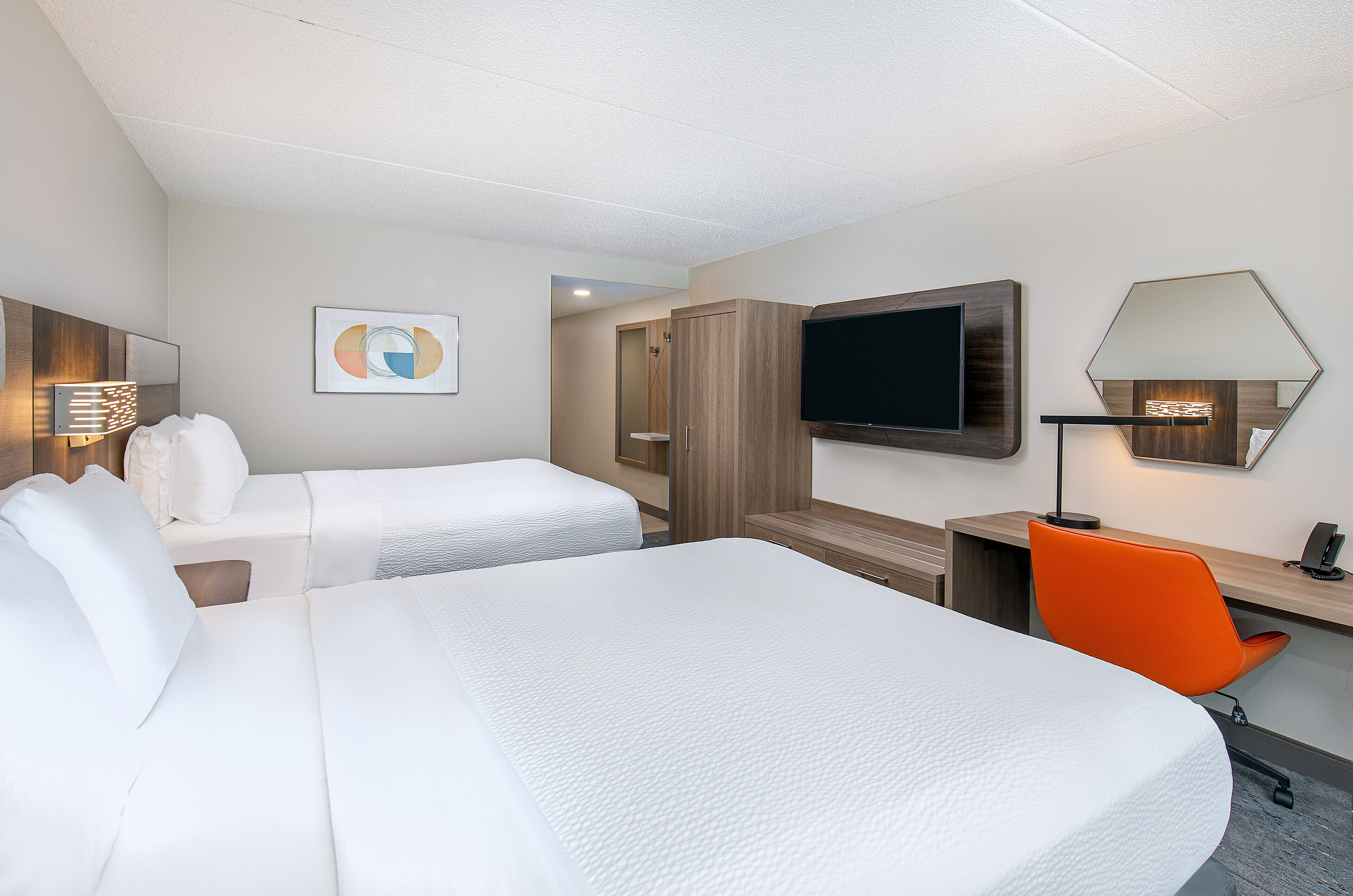 Une chambre d’hôtel avec deux lits, un bureau, une chaise, une télévision, un miroir et une œuvre d’art accrochée au mur.