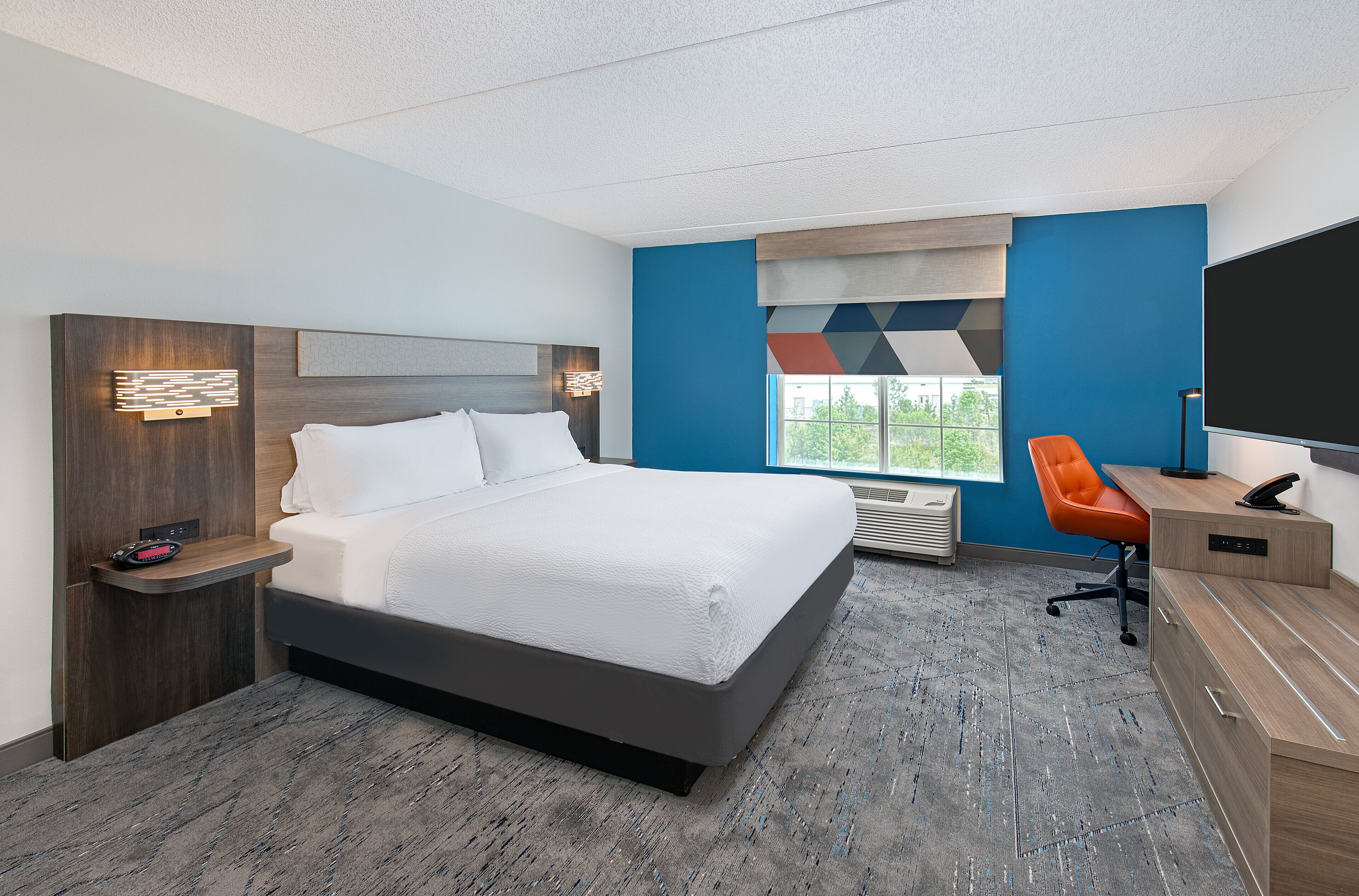 A hotel room with a bed, bedside table, desk, chair, and a large window.
