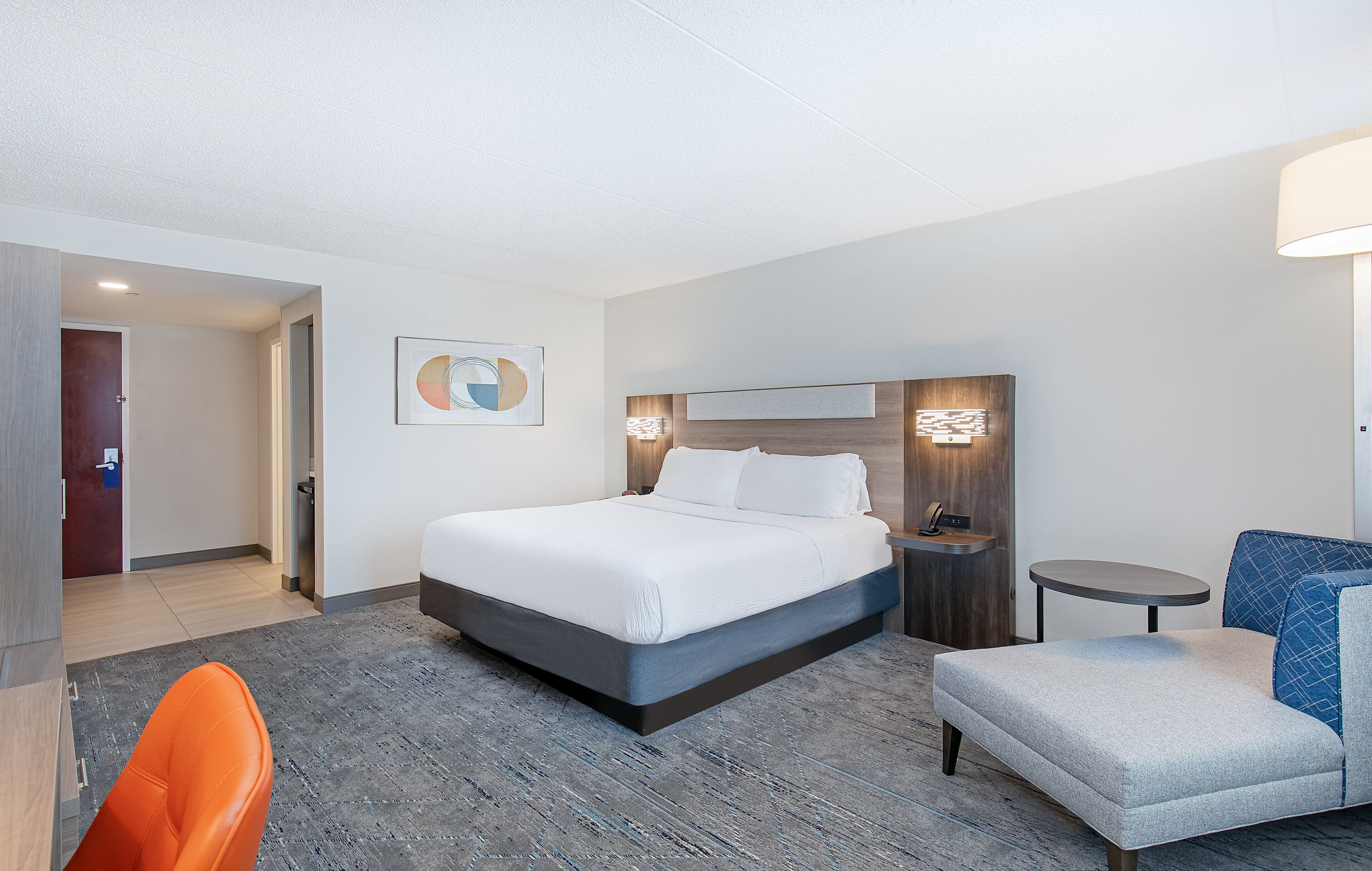 A hotel room with a bed, a desk, a chair, and a bedside table.