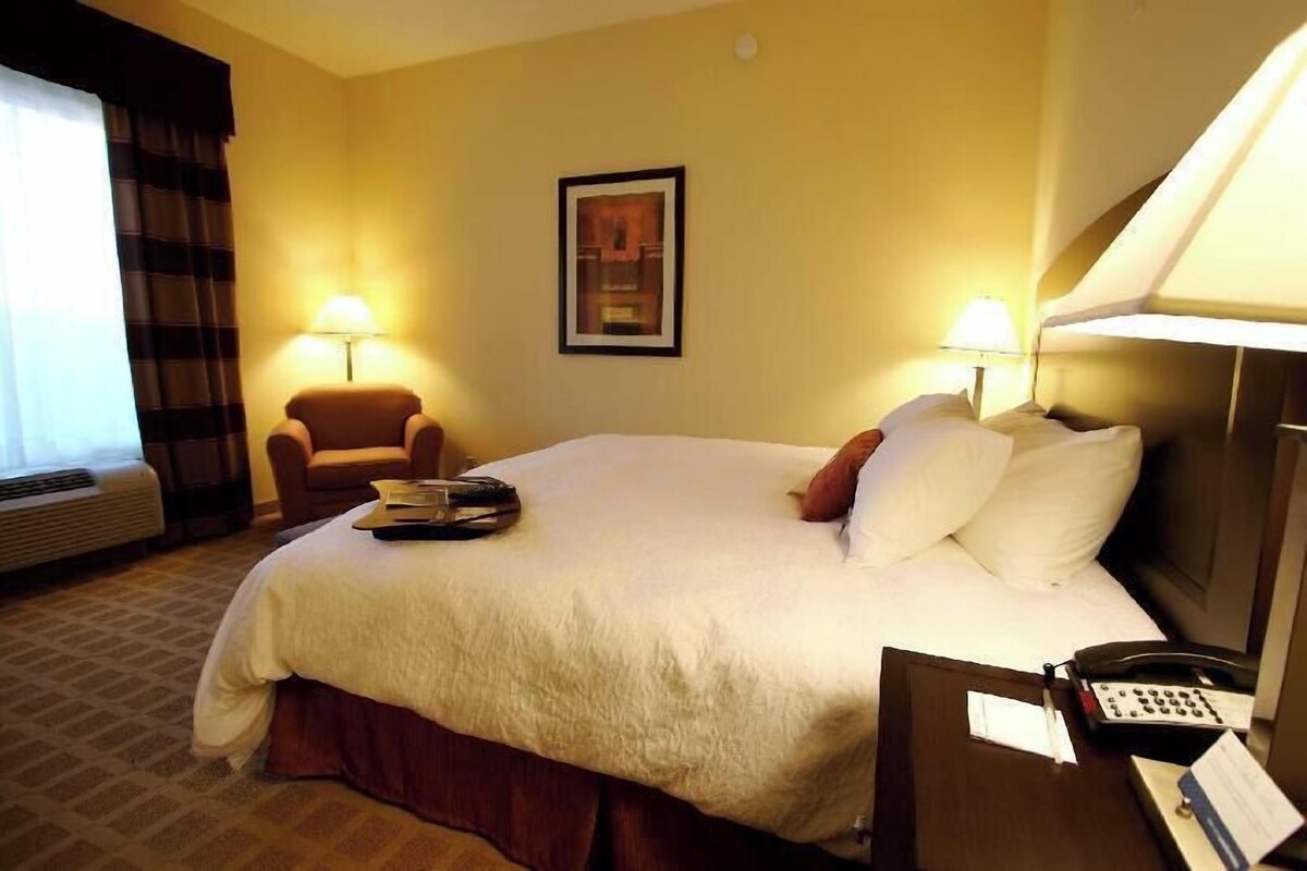 A hotel room with a bed, two bedside lamps, a chair, a desk with a phone, and a framed picture on the wall.