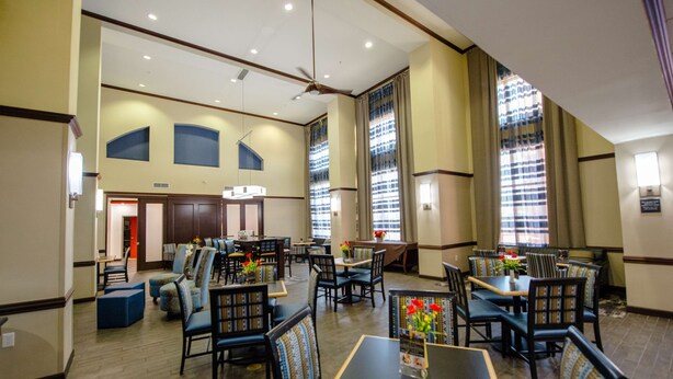 A spacious hotel dining area with multiple tables and chairs, large windows with curtains, and a ceiling fan. - Hampton Inn & Suites Port Arthur (Port Arthur)