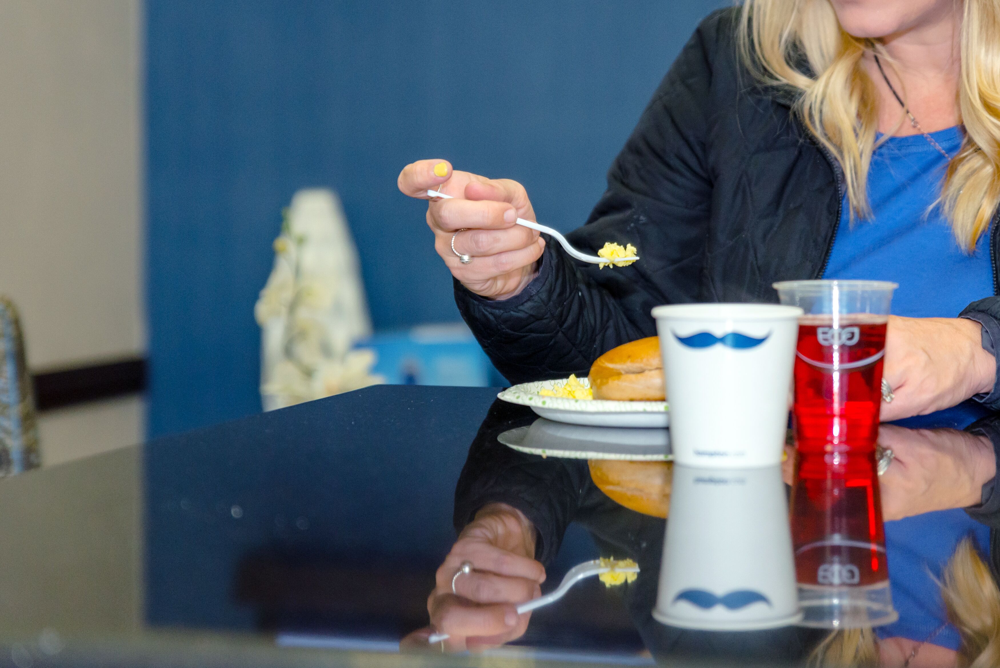Una persona sentada a una mesa con un plato de comida, una bebida en un vaso alto y una taza blanca con un diseño azul.
