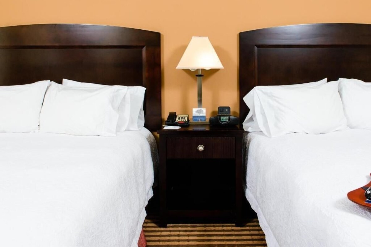A hotel room with two beds, a nightstand with a lamp, a phone, and a clock.