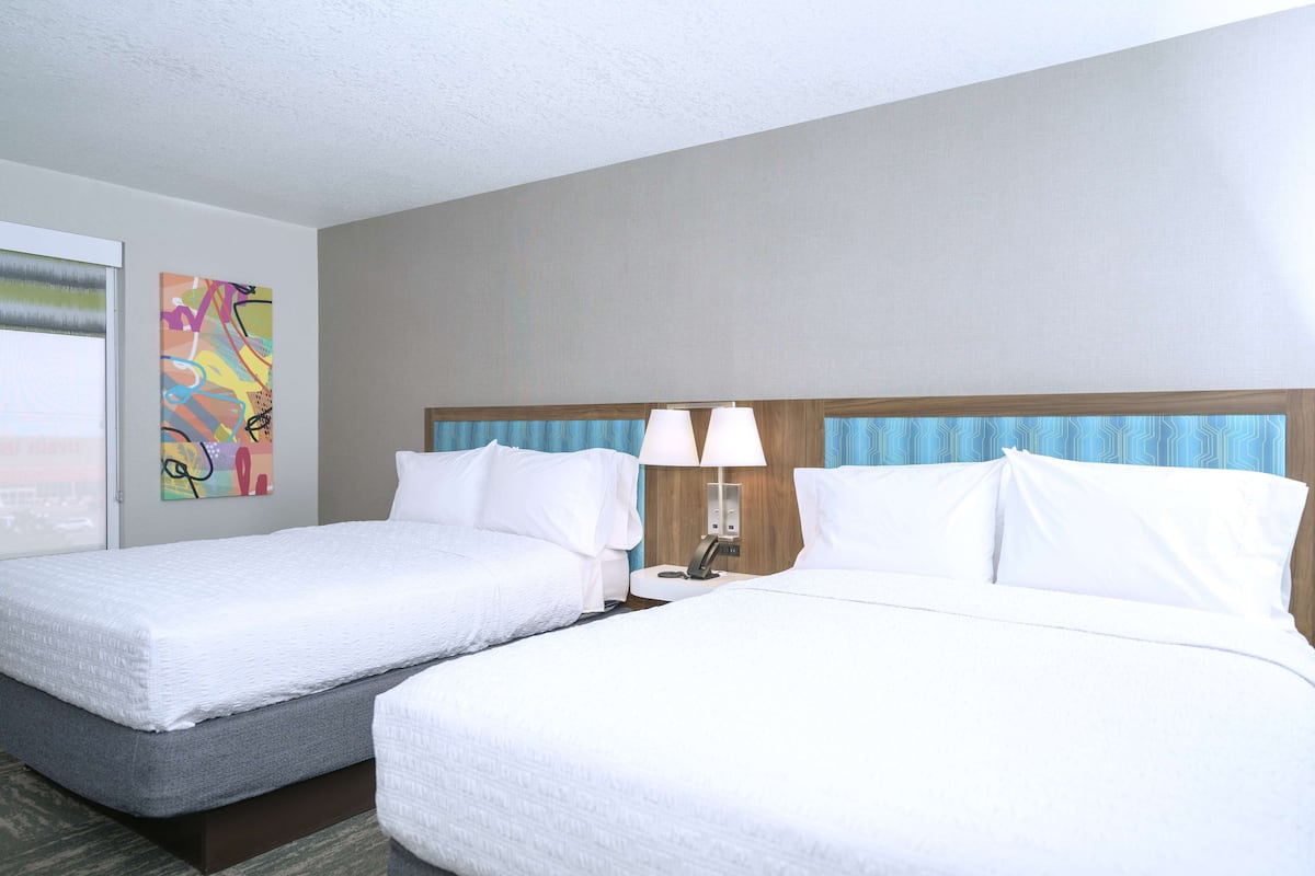 A hotel room with two beds, a nightstand with a lamp, and a colorful abstract painting on the wall.