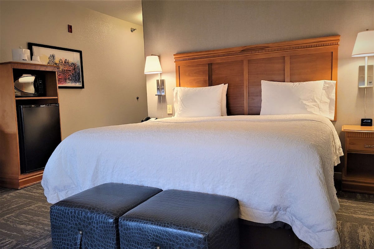 A hotel room with a large bed, a wooden headboard, a small desk, a lamp, and a refrigerator.