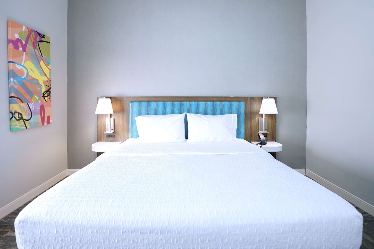 A hotel room with a large bed, two bedside lamps, a wooden headboard, and a colorful abstract painting on the wall.
