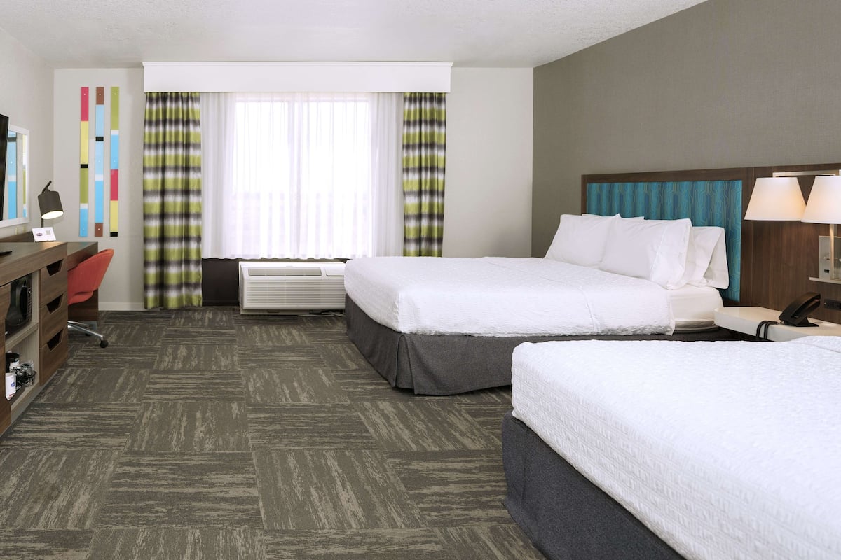 A hotel room with two beds, a desk, a chair, a desk lamp, a telephone, and a window with curtains.