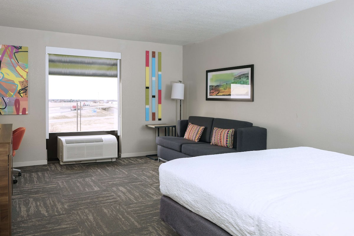 A hotel room with a bed, a sofa, a window with a view, a radiator, and a painting on the wall.