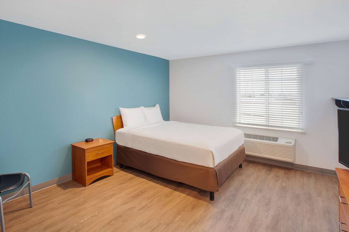 A bedroom with a bed, a nightstand, a chair, and a window with blinds.