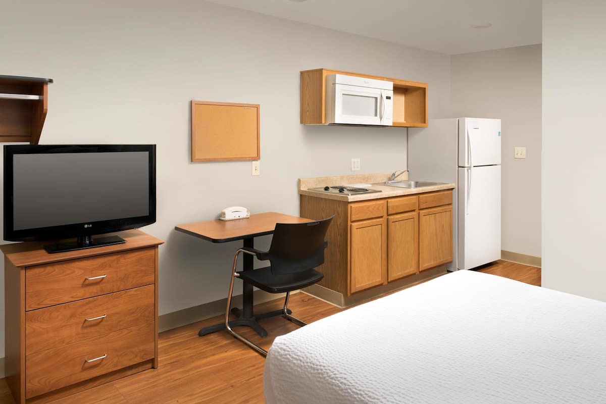 A hotel room with a bed, a desk with a chair, a television, a microwave, a refrigerator, and wooden cabinets.
