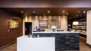 Un espace d’accueil d’hôtel au design moderne, comprenant un comptoir d  ’accueil, un panneau d’affichage et un bar avec des étagères remplies de bouteilles.