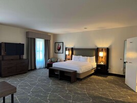 Quarto de hotel com uma cama grande, televisão em uma cômoda, escrivaninha, cadeira e uma porta.