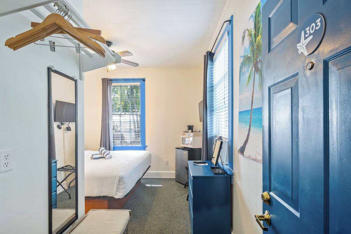 Une chambre d’hôtel équipée d’un lit, d’un bureau, d’un miroir, d’une armoire et d’une porte avec une plaque numérotée.