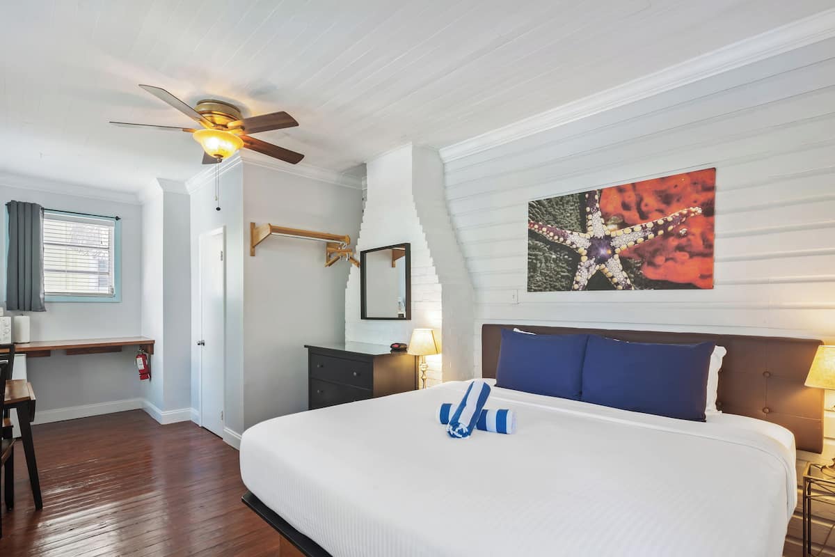 Une chambre d’hôtel avec un grand lit, un ventilateur de plafond, un bureau avec une chaise, une table de chevet avec une lampe et un tableau au mur.