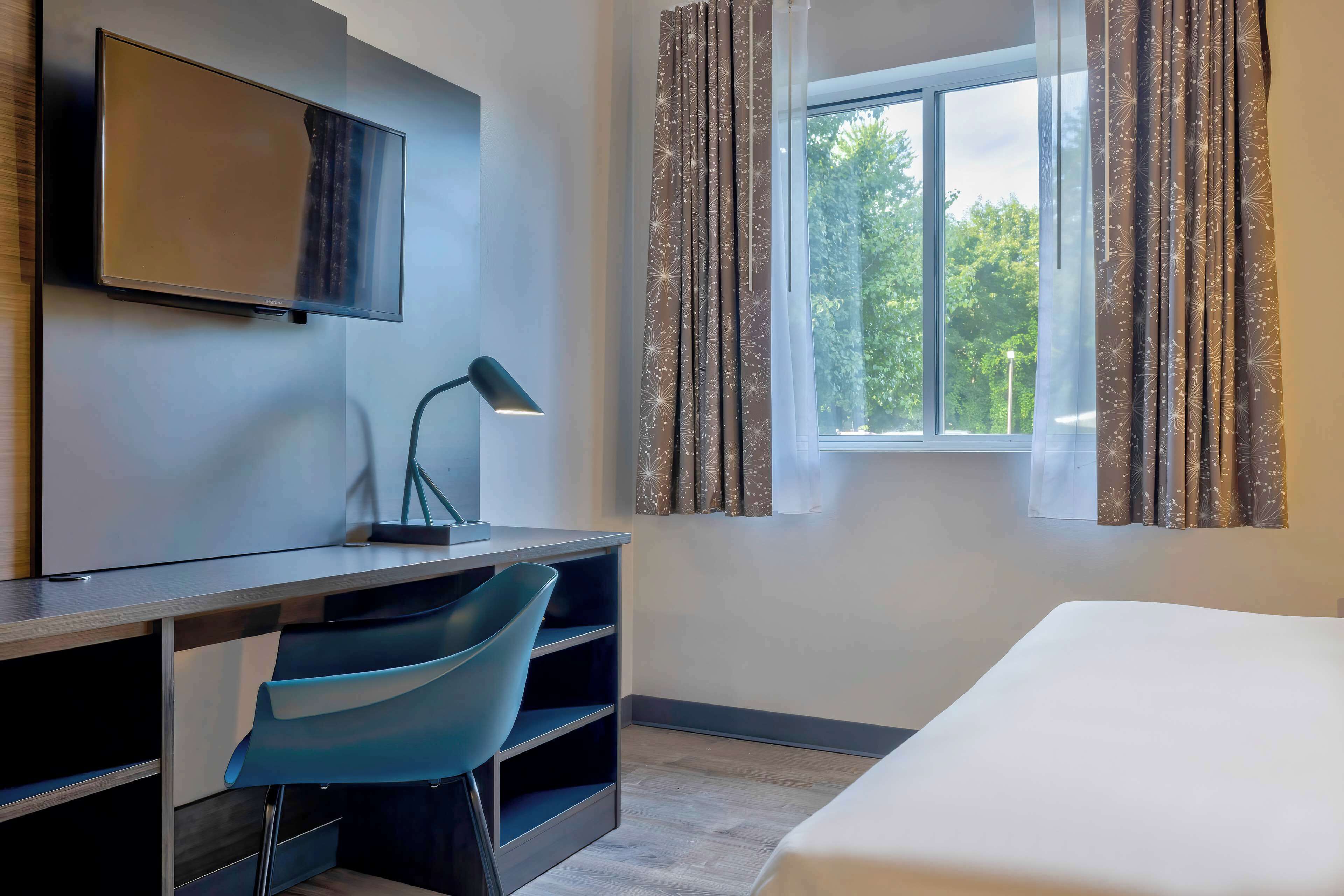 Ein Hotelzimmer mit einem Bett, einem Schreibtisch mit Stuhl, einem an der Wand befestigten Fernseher und einem Fenster mit Vorhängen.