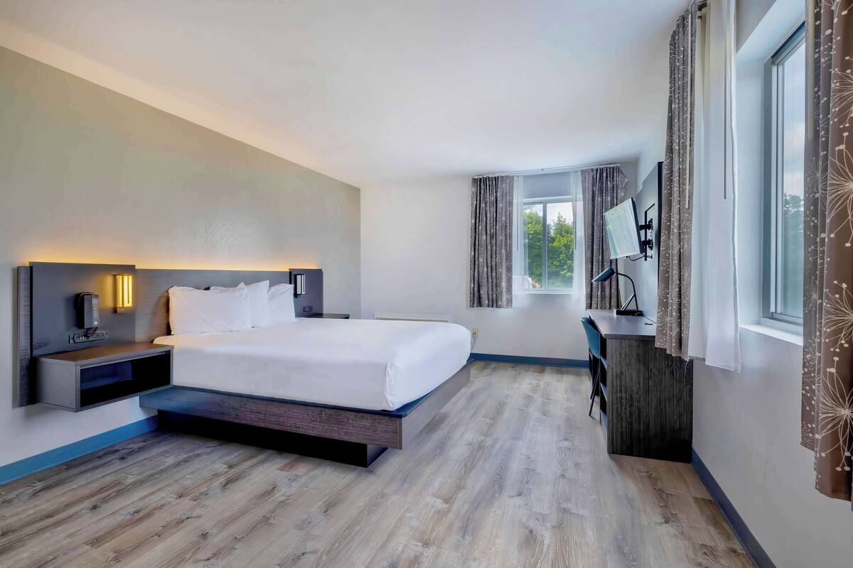 A modern hotel room with a bed, bedside table, lamp, and a desk with a chair.