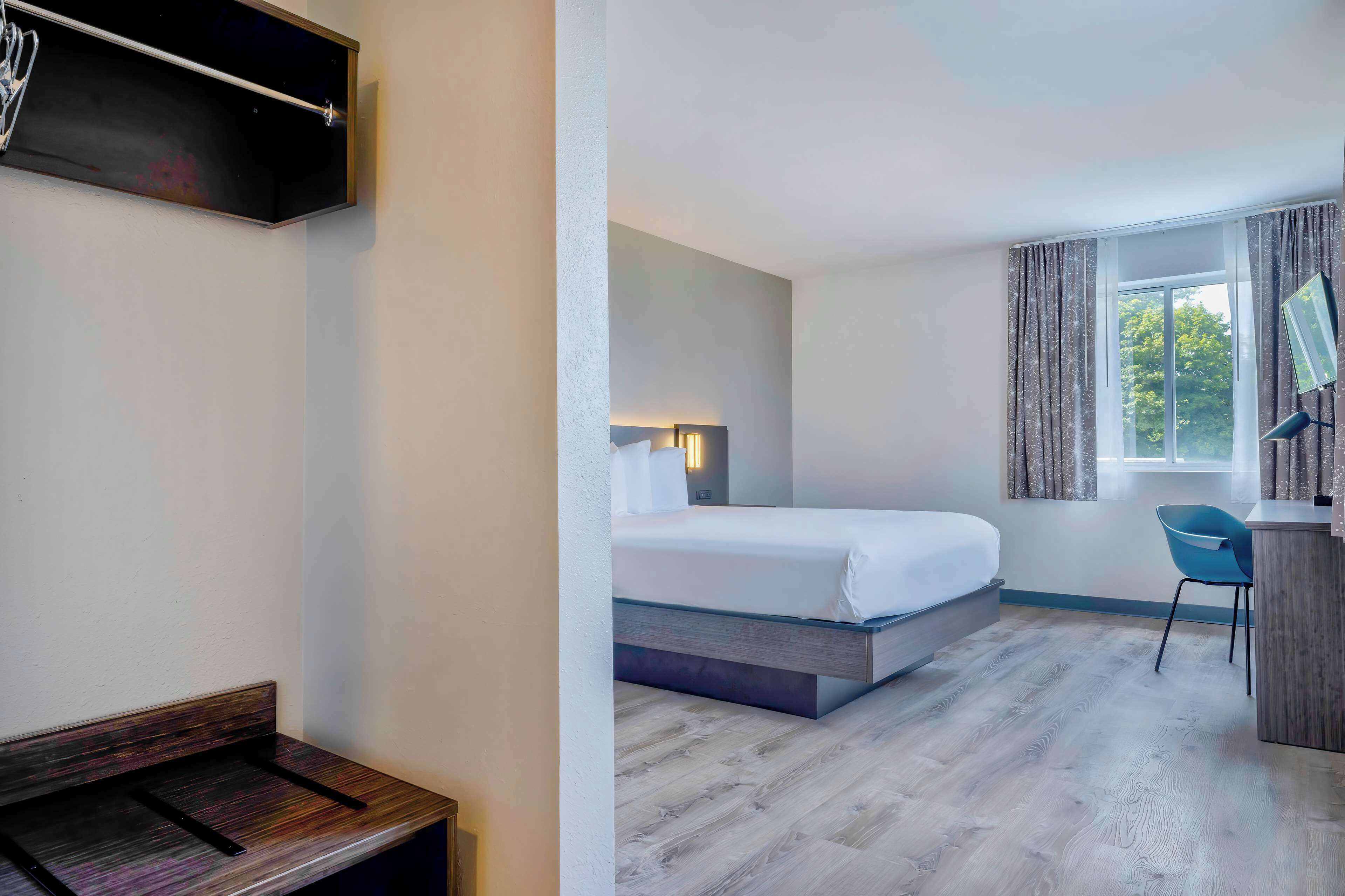 A hotel room with a bed, a desk, a chair, a TV mounted on the wall, and a window with curtains.