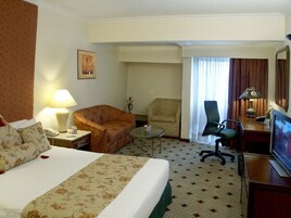 A hotel room with a large bed, a desk with a television, a chair, a small table, and a lamp.