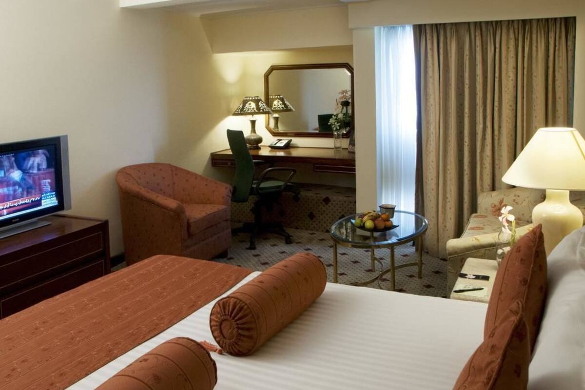 Habitación de hotel con cama, escritorio, silla, televisión y una mesita con fruta.