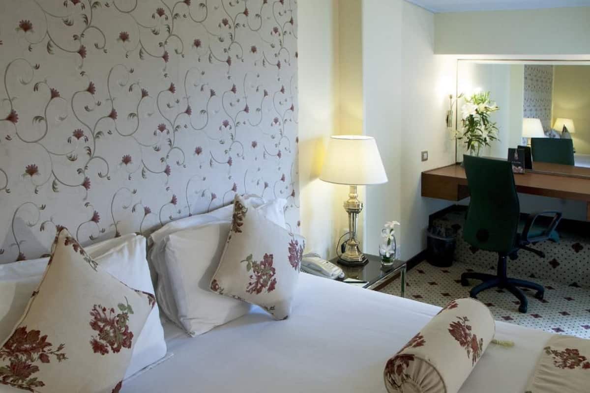 Habitación de hotel con cama, almohadas estampadas con motivos florales, escritorio con lámpara y una silla.