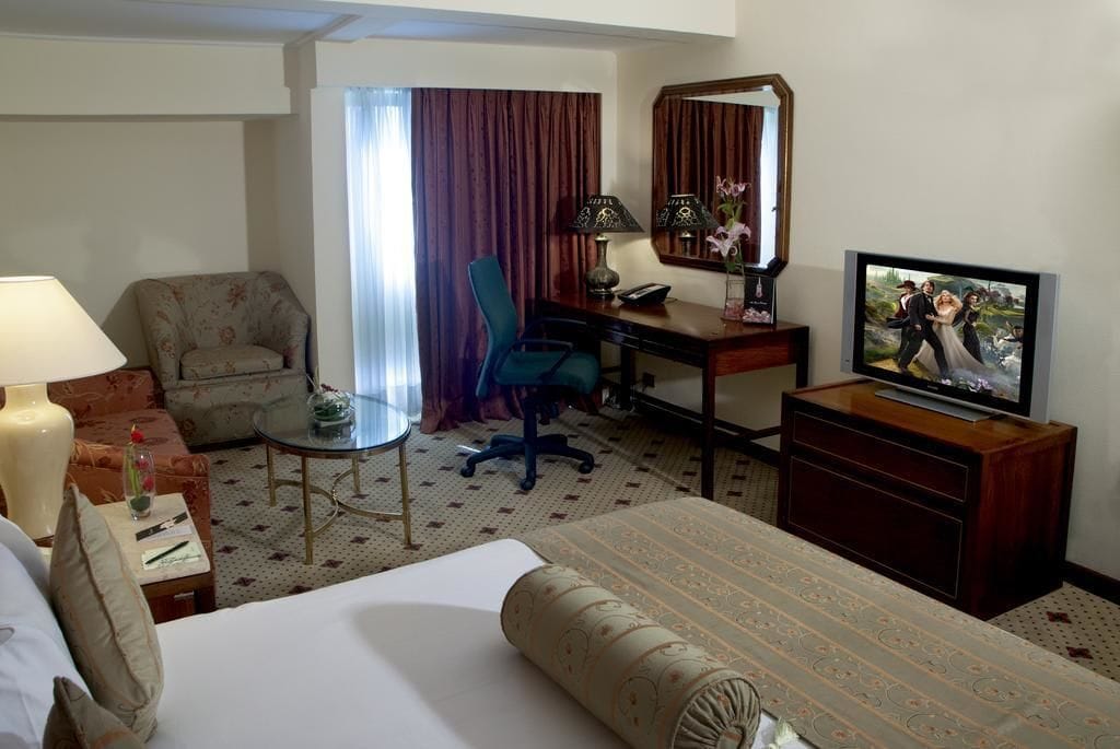 A hotel room with a bed, a desk, a chair, a television, and a small table with a lamp.
