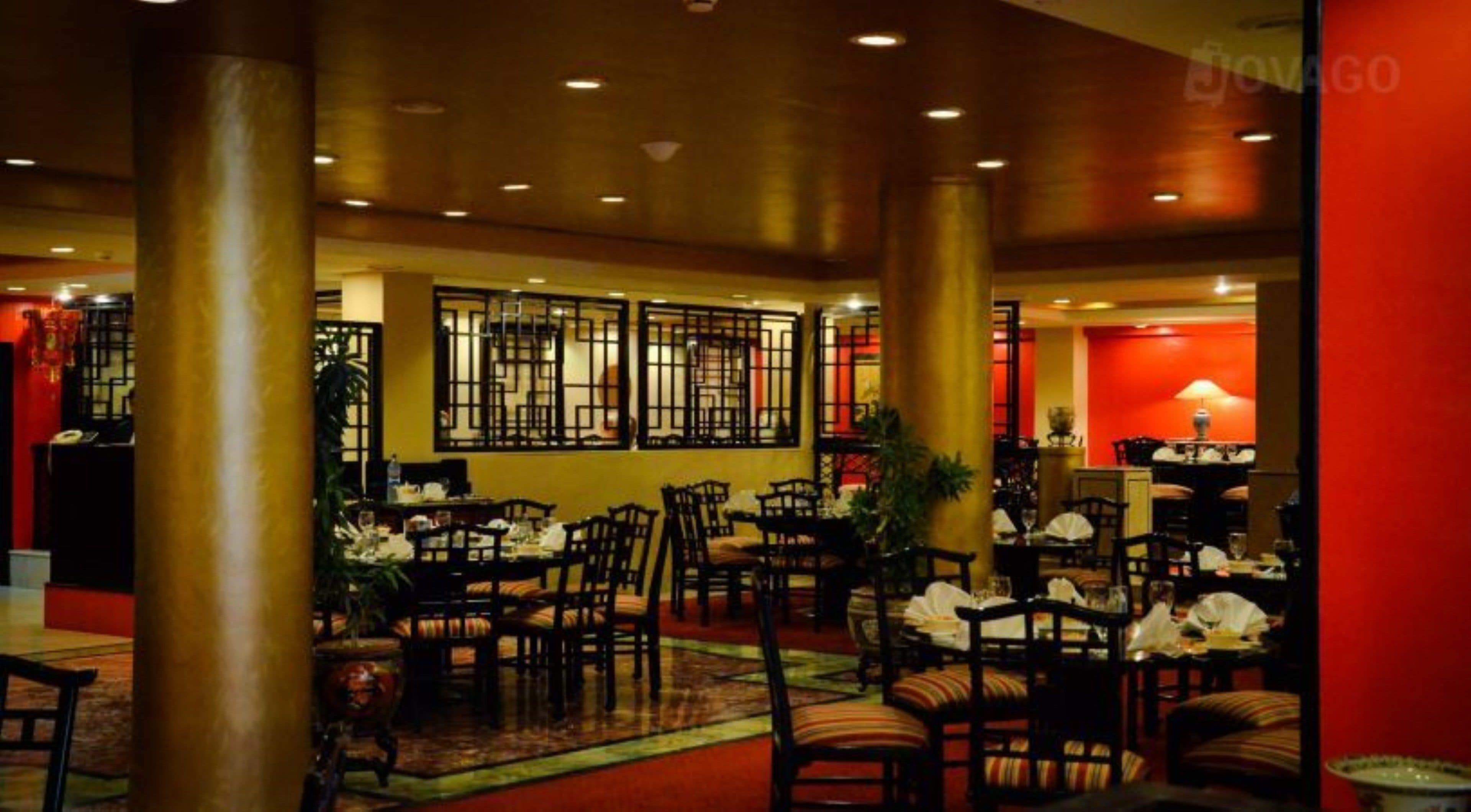 Un hall de hotel con varias mesas y sillas para comer, un mostrador de recepción y una pared decorativa con un diseño geométrico.