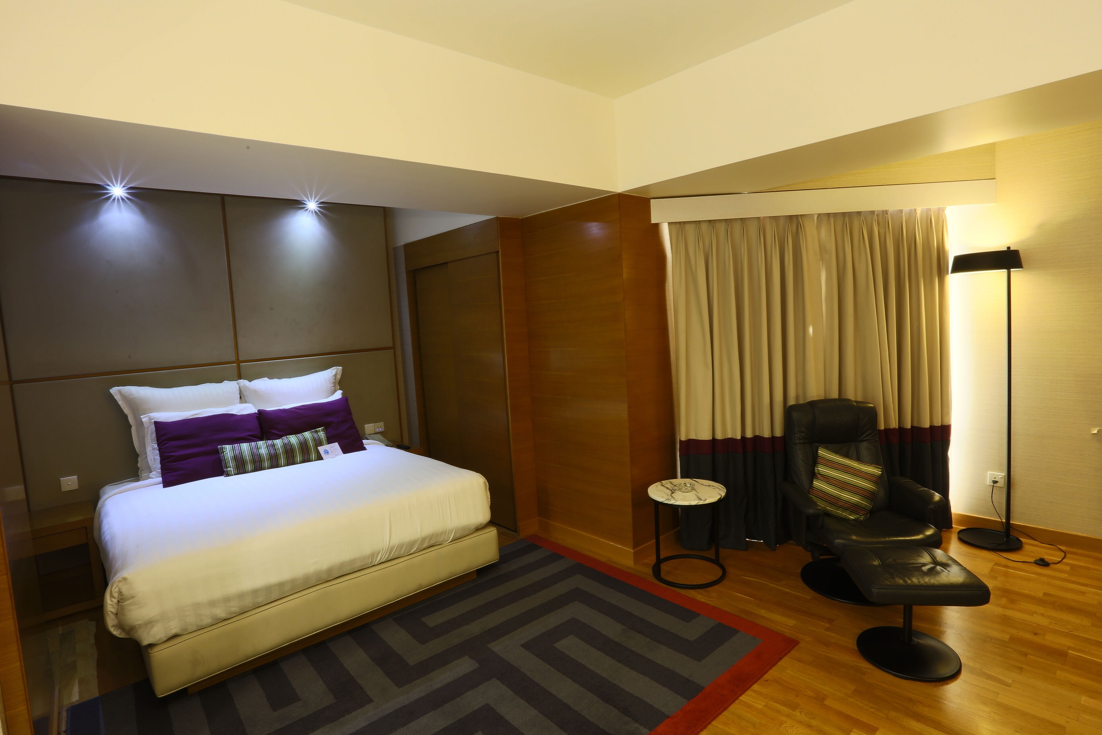 A modern hotel room with a large bed, a small side table, a chair, and a floor lamp.
