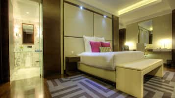 A modern hotel room with a large bed, bedside tables, a bench, a bathroom with a glass shower enclosure, and a large mirror.