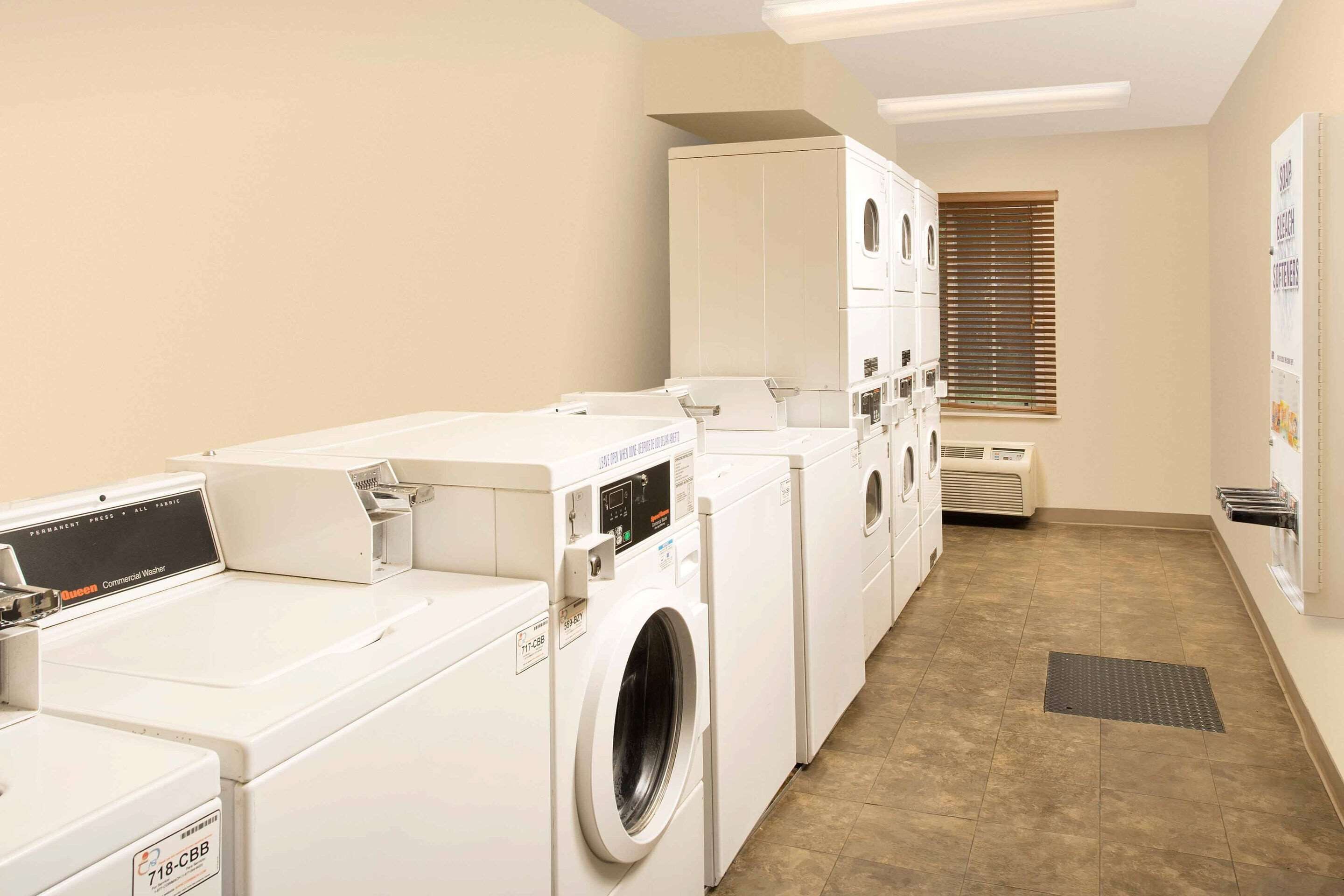 A laundry room with multiple washing machines and dryers, a ventilation unit, and a window with blinds.