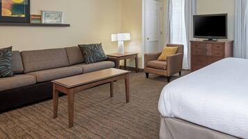 A hotel room with a sofa, armchair, small table, lamp, TV, and a bed.