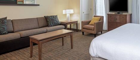 A hotel room with a sofa, armchair, small table, lamp, TV, and a bed.