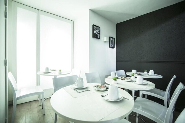 A modern dining area with white tables and chairs, a large window, and framed artwork on the wall. - Hotel Schtak (Cannes)