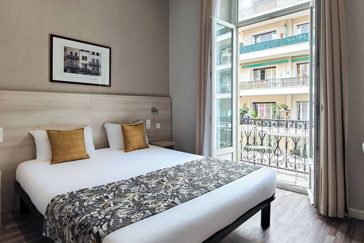 A hotel room with a large bed, a framed picture on the wall, and a balcony with a view of other buildings.
