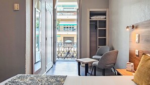 A narrow room with a bed, a small table, two chairs, a wooden headboard, and a view of a balcony with a railing. - Hôtel Helvétique (Nice)