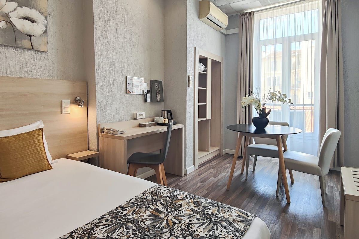 A hotel room with a bed, desk, chair, and a small table with a vase of flowers.