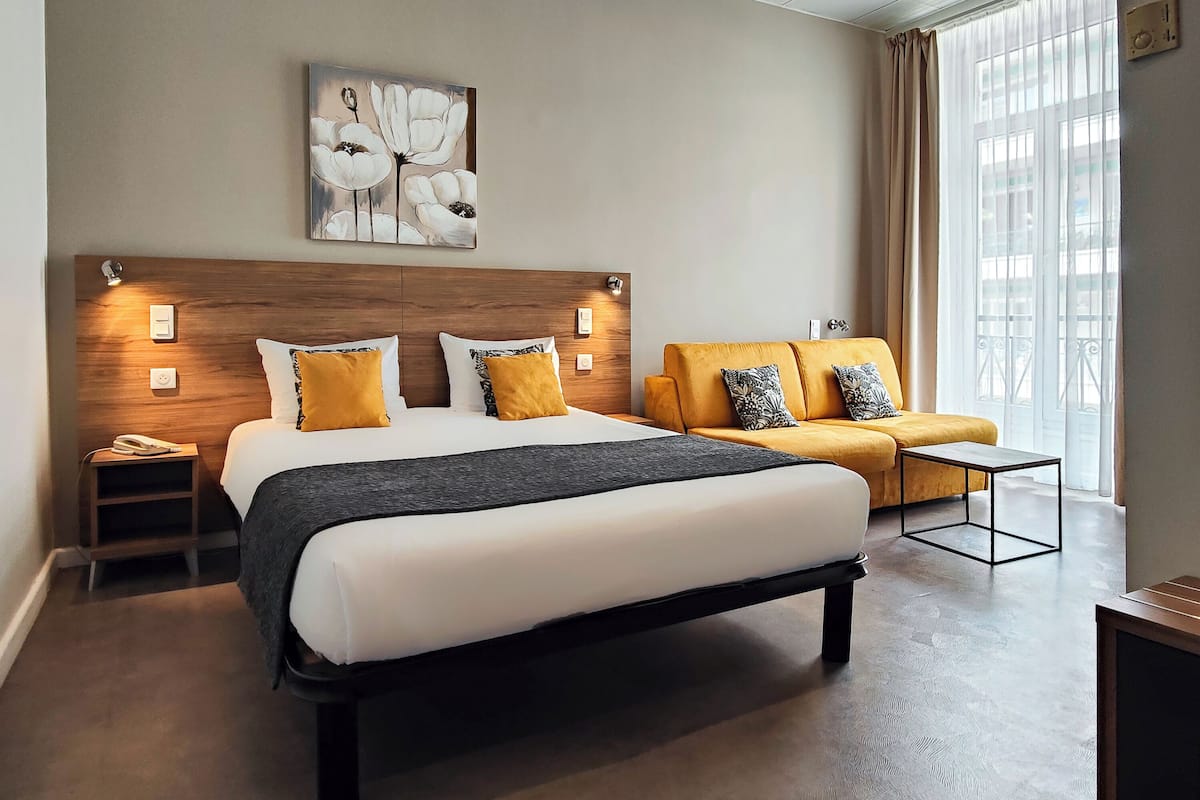 A modern hotel room with a large bed, a sofa, a bedside table, a nightstand, and a painting on the wall.