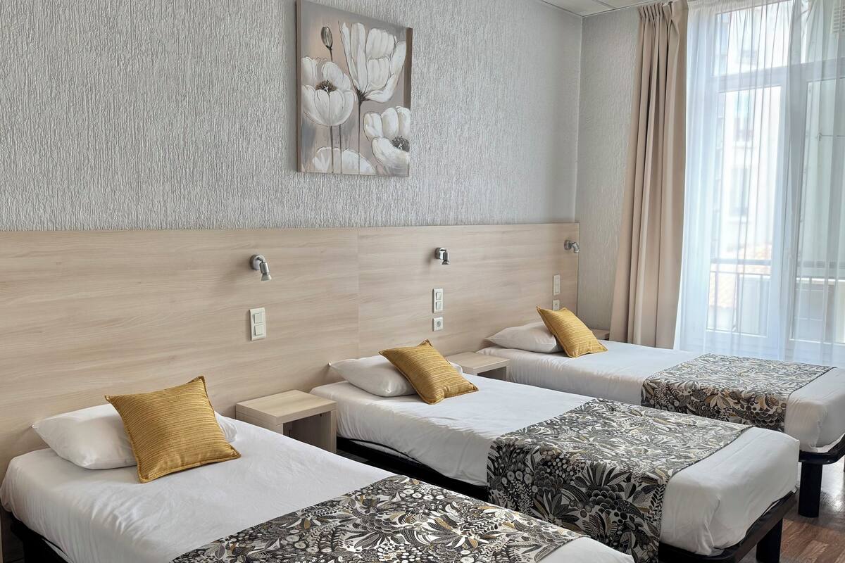A hotel room with three beds, each with a patterned bedspread and a single pillow. There is a wall-mounted artwork above the beds, and the room has a wooden headboard.
