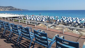 A seaside view with rows of blue chairs and white umbrellas, a railing, and a clear blue sky. - Hôtel Helvétique (Nice)
