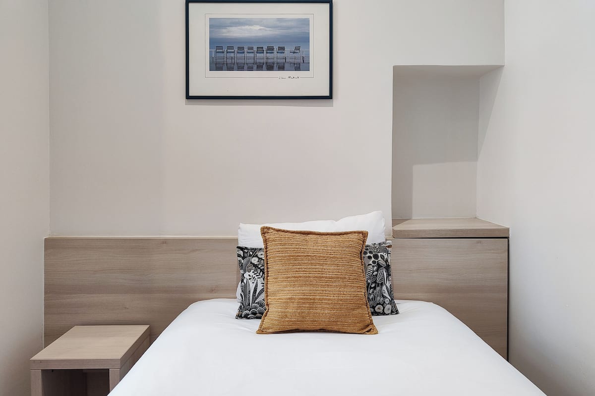 A neatly arranged hotel room with a bed, pillows, a bedside table, a nightstand, and a framed picture on the wall.