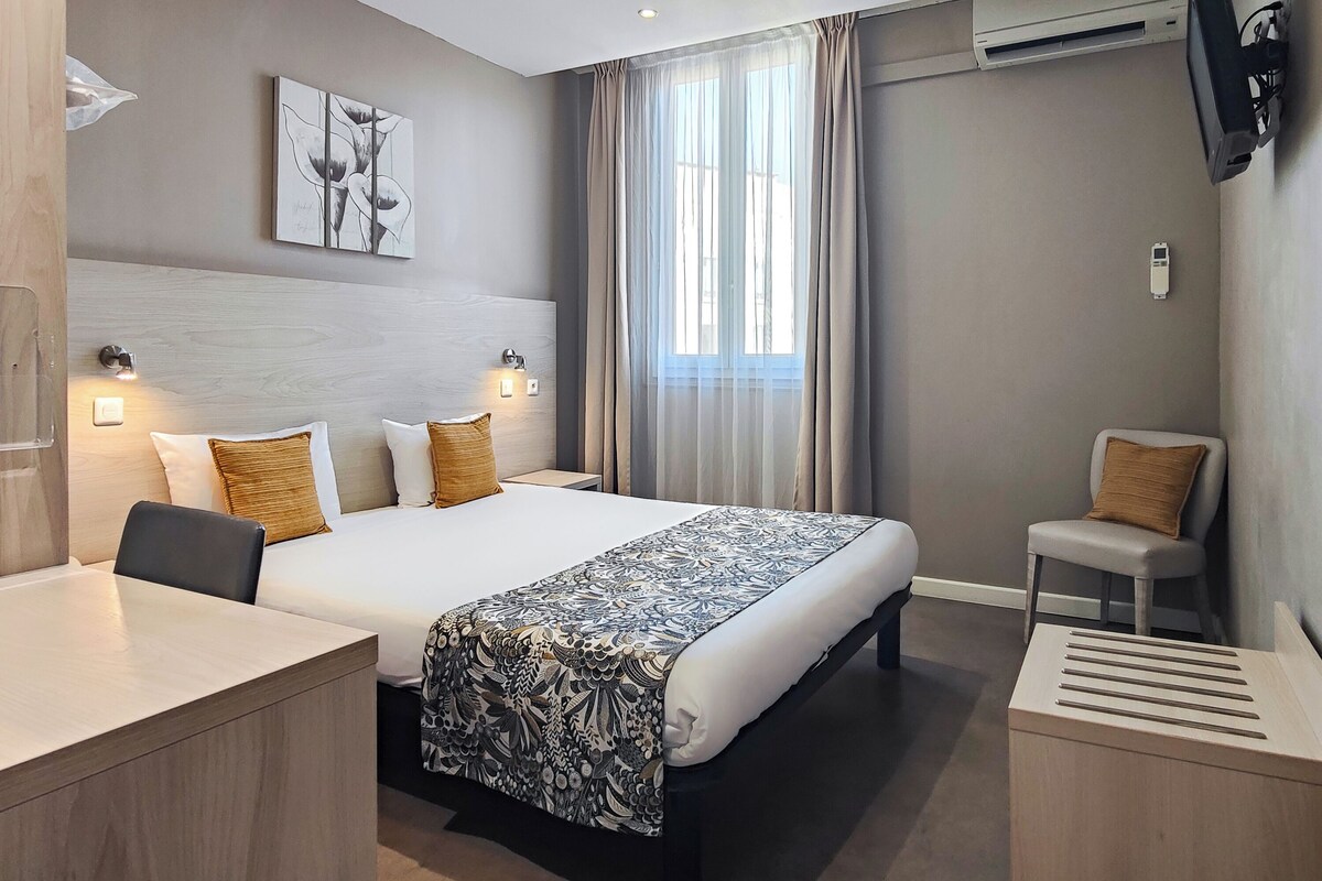 A hotel room with a bed, bedside tables, a desk, a chair, a TV, and a window with curtains.